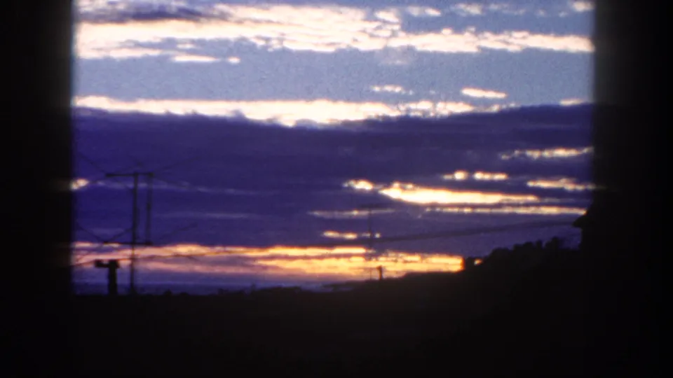 1962 San Diego frame with golden sunrise light under layered clouds, framed by silhouetted poles, rooftops, and hillside.