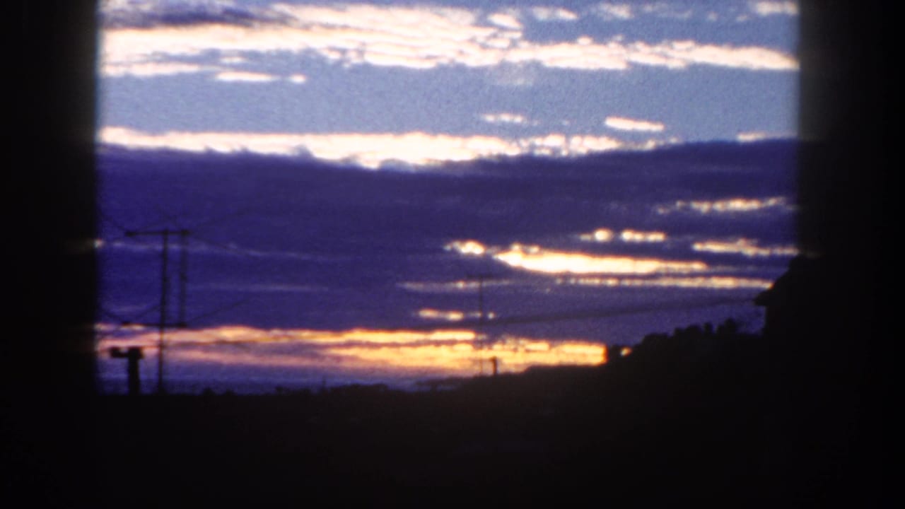 1962 San Diego 8mm frame showing blue-purple clouds and warm horizon light above silhouetted poles, wires, and a rooftop edge.