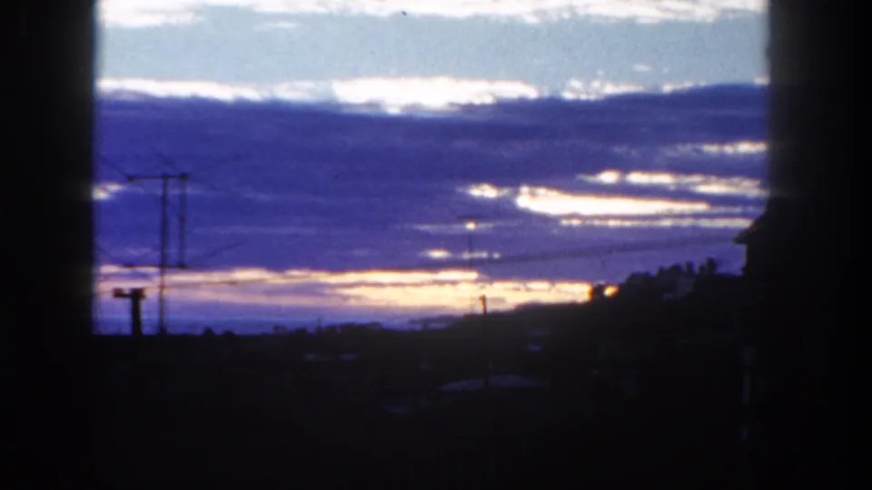 1962 San Diego 8mm frame showing a brightening or fading horizon behind cloud bands, wires, antenna, and roofline silhouettes.