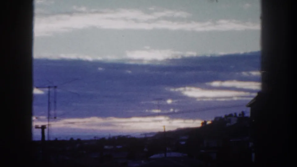 8mm frame from San Diego in 1962 showing cloud bands, a darkened small-town horizon, utility poles, wires, and window-like framing.