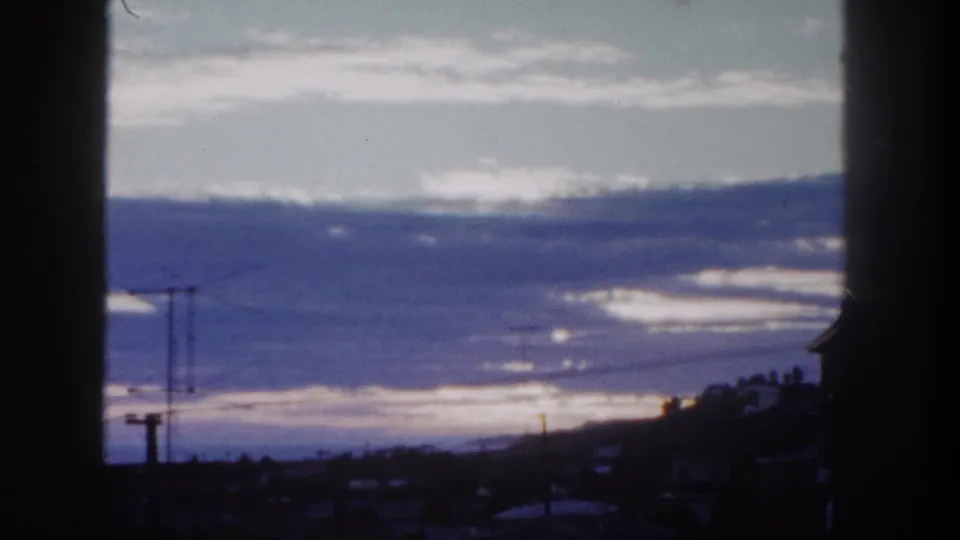 1962 San Diego 8mm frame showing a sunrise or sunset sky with silhouetted utility lines, low rooftops, hillside forms, and dark foreground borders.