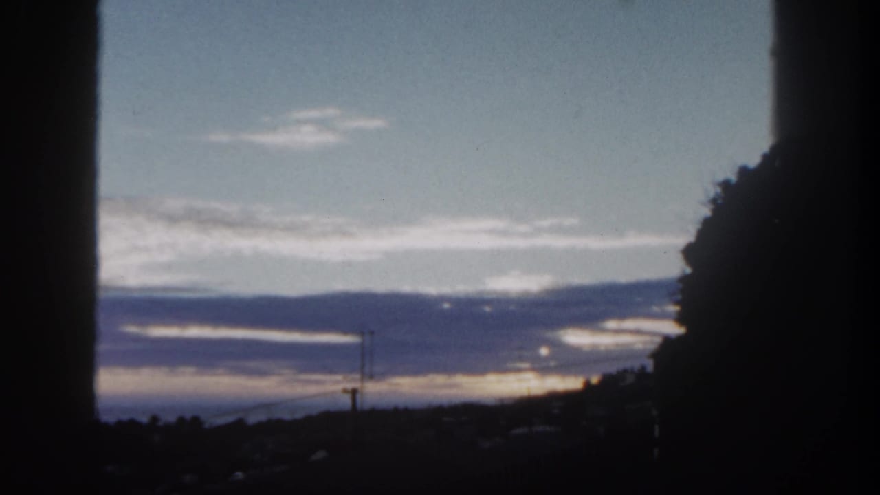 Sunset over distant hills and clouds, framed by dark vertical edges and a tree silhouette.