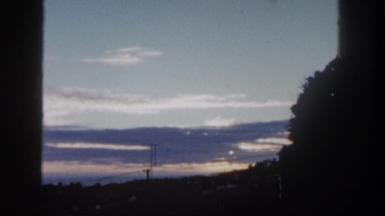 1962 San Diego home-movie frame with sunset clouds over hills, seen through dark foreground shapes and a large tree silhouette.