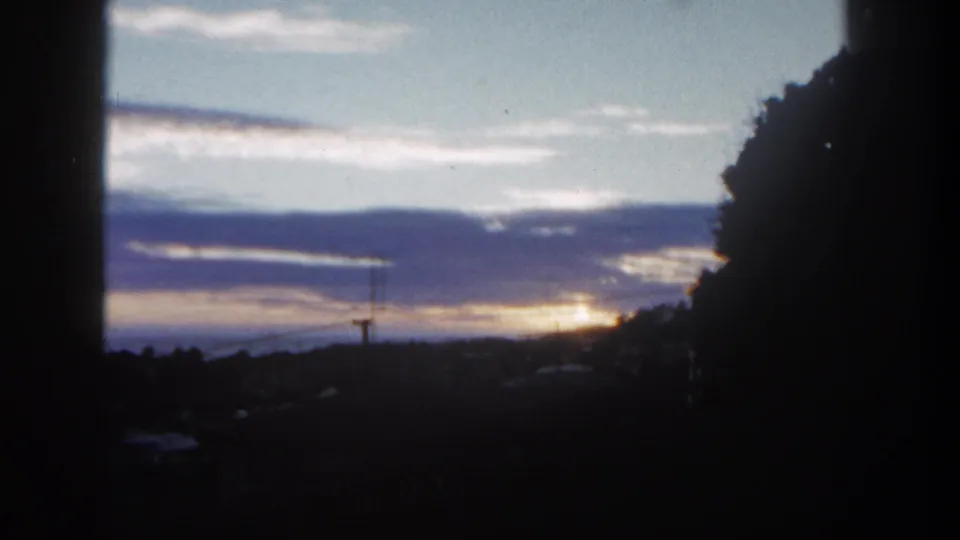 1962 San Diego dusk view with layered clouds, a bright low sun glow, dark trees, and faint horizon structures.