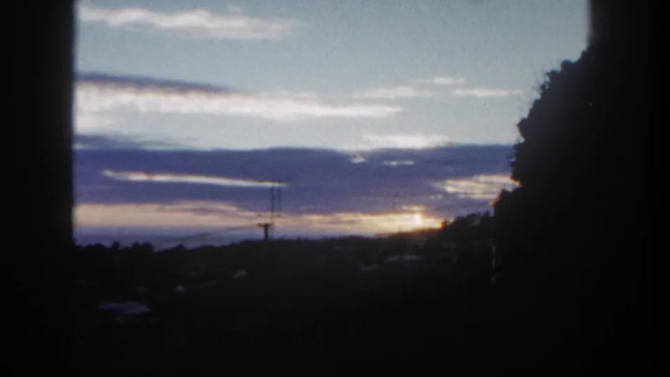 San Diego 1962 home-movie frame with a dim foreground, silhouetted trees, purple clouds, and a faint utility structure near the horizon.