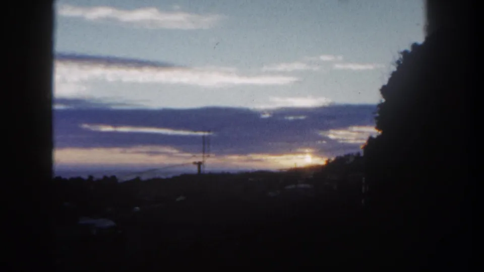 1962 San Diego sunset frame with blue-purple cloud bands, a bright horizon glow, dark trees at right, and low landscape silhouettes.