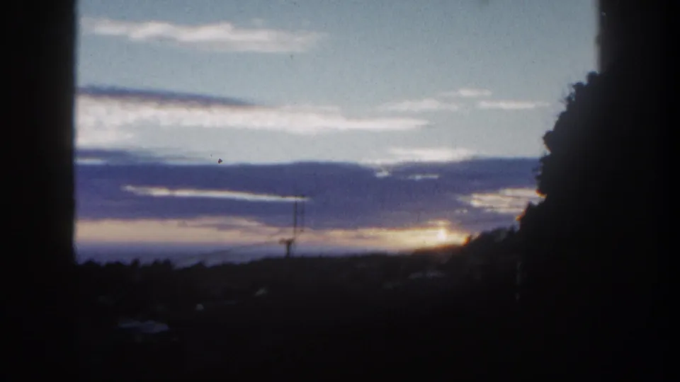 1962 San Diego home-movie frame showing a low sun through layered clouds over a dark suburban horizon.