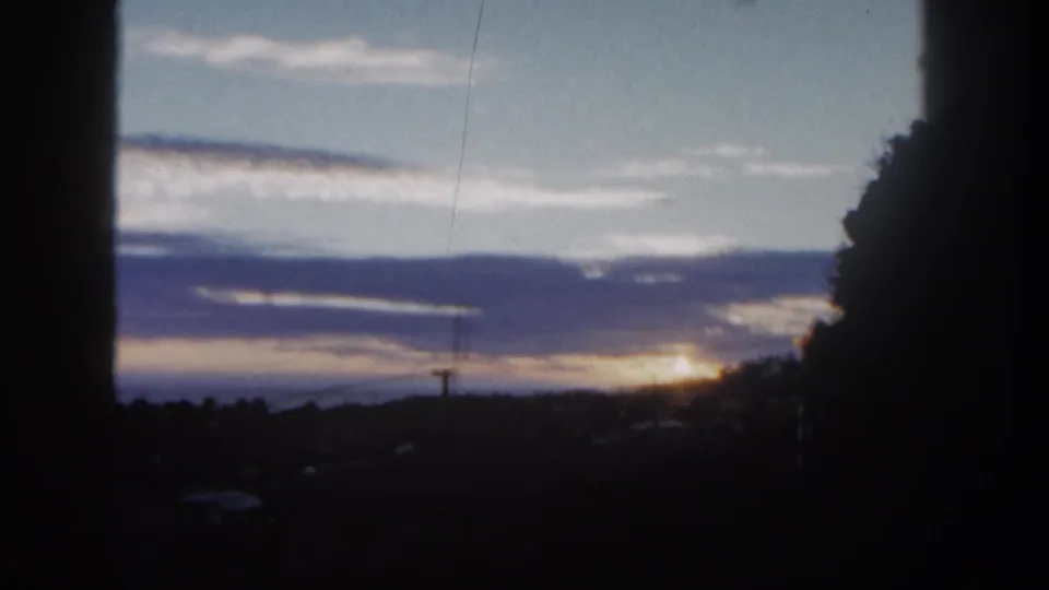 1962 San Diego home-movie frame of a sunlit horizon behind clouds, with trees and a dark window-like edge.