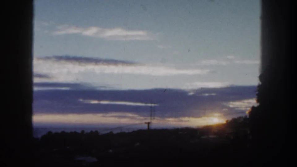 1962 San Diego sunset frame with the sun at the right horizon, layered clouds, silhouetted trees, and faint distant structures.
