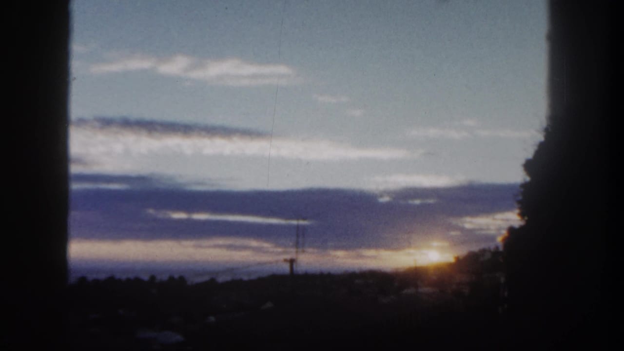 San Diego 1962 home-movie frame showing a low sunrise, silhouetted hillside, distant tower, and layered blue-gray clouds.