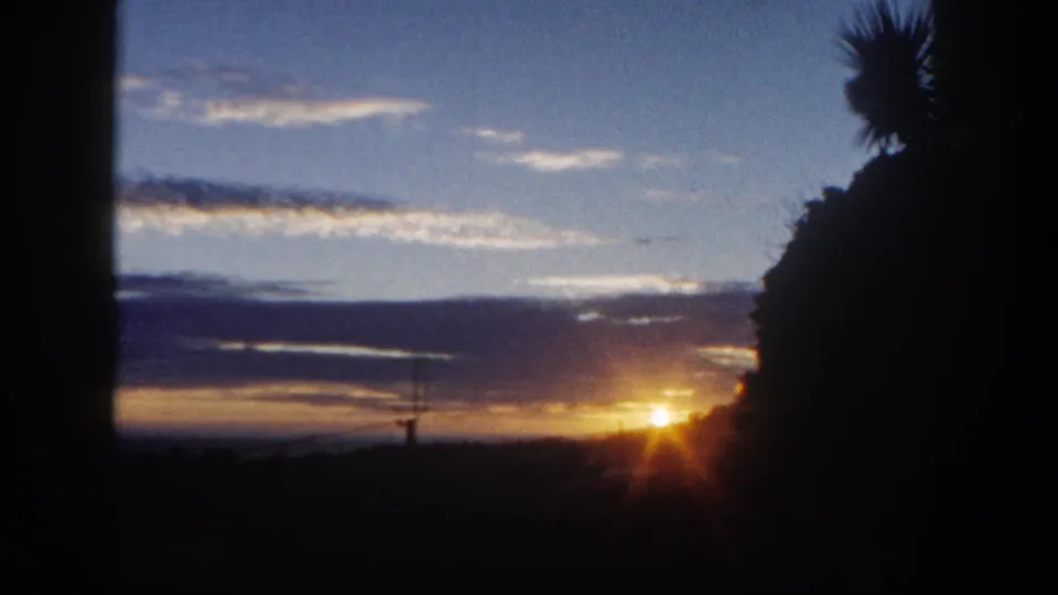 1962 San Diego 8mm frame with bright sunrise flare, dark hillside, palm silhouette, and blue-purple cloud layers.