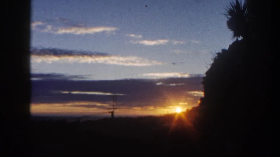 1962 San Diego home-movie frame showing a low orange sun near the horizon, blue clouded sky, and dark tree silhouettes.