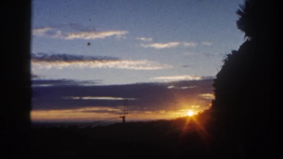 1962 San Diego home-movie frame of an orange sunset under blue clouded sky, framed by dark trees and palm foliage.