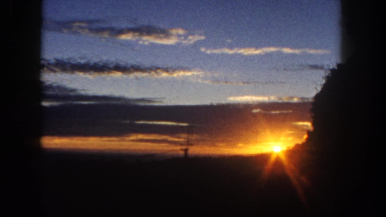 1962 San Diego-area 8mm frame with a bright sun flare at the horizon, layered clouds, blue evening sky, and silhouetted trees.