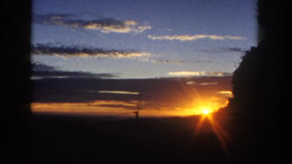 1962 San Diego-area 8mm frame showing a low orange sun partly softened by exposure bloom beneath blue sky and wispy clouds.