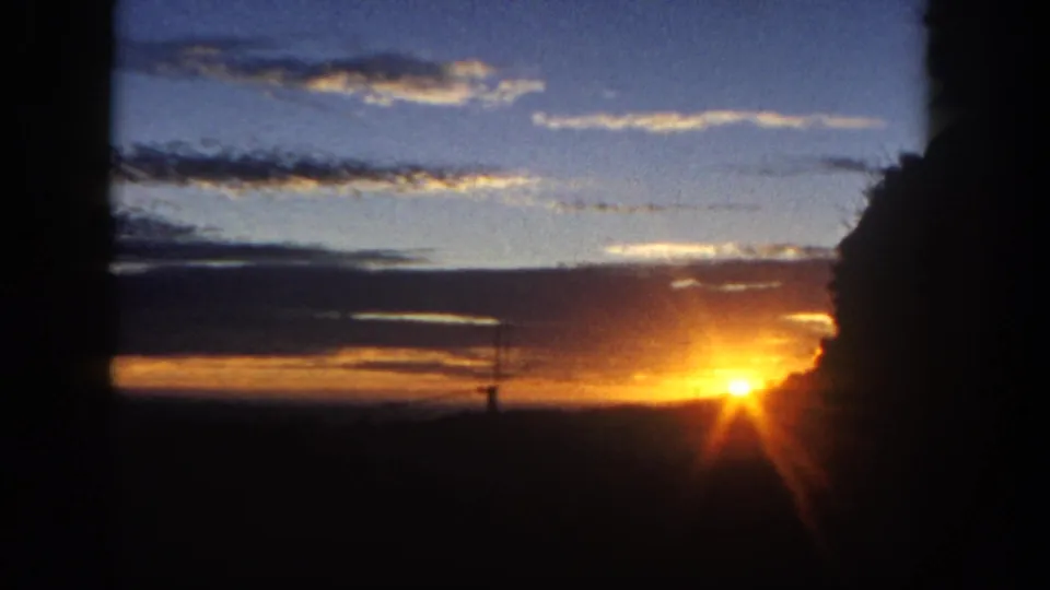 1962 San Diego-area 8mm frame of a star-like sun flare near the horizon, framed by dark tree edges and layered evening clouds.