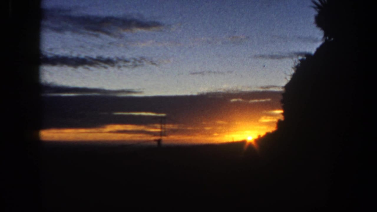 1962 San Diego 8mm frame with a low sunset glow, blue dusk sky, layered clouds, a silhouetted antenna or tower, and dark foliage.