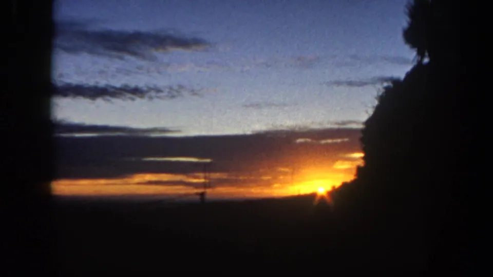 1962 San Diego 8mm frame showing the sun lower at the edge of the horizon, with blue sky, orange clouds, and dense silhouetted foliage.