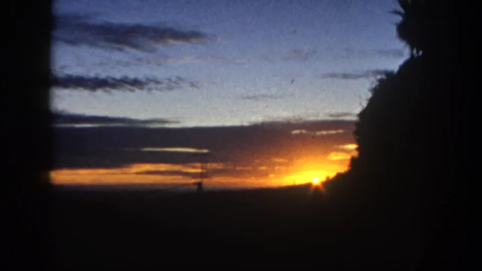 1962 San Diego 8mm frame with the sunset partly obscured, layered dusk clouds, a silhouetted antenna or tower, and dark foliage at right.