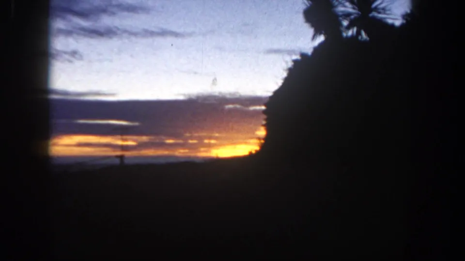 1962 San Diego 8mm frame showing a low orange sunset, clouds, and silhouetted foreground foliage.