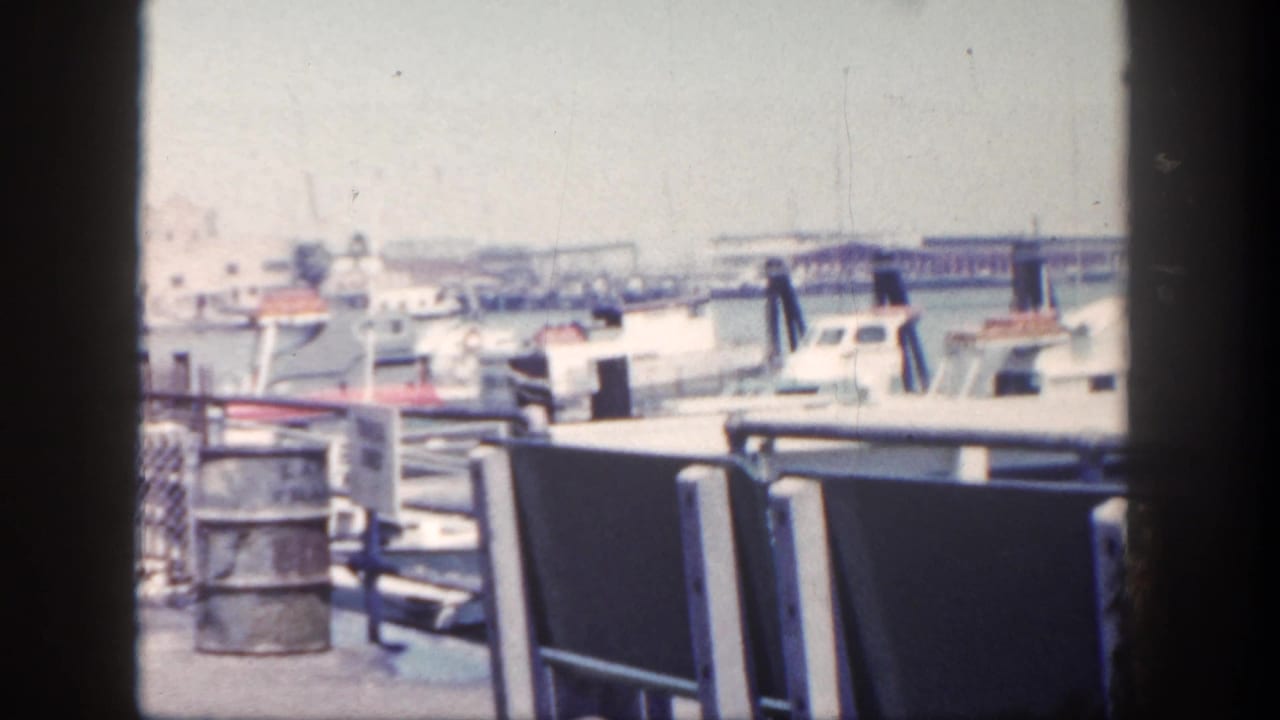 1962 San Diego waterfront scene showing dock railings, moored boats, pilings, hazy water, and dark vertical framing at the edges.
