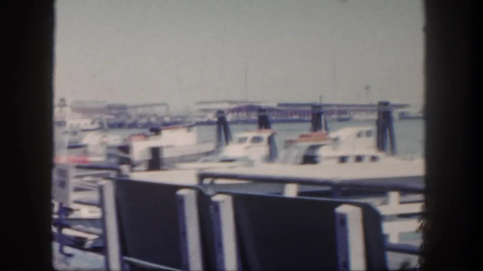1962 San Diego marina footage with foreground railing, docked cabin boats, pilings, water, and a distant waterfront structure.