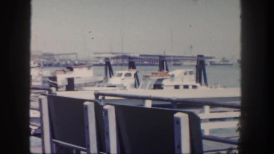 1962 San Diego marina view of moored boats, railings, pilings, calm water, and a distant pier or bridge-like structure.