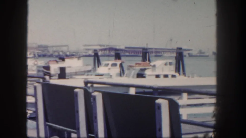 1962 San Diego harbor view with white boats, dark pilings, pier railings, and benches under a hazy sky.