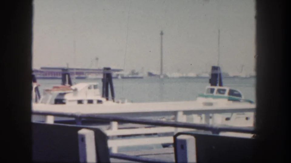 1962 San Diego harbor shot with a white boat, foreground railing, dark pilings, and a tall distant tower-like structure.