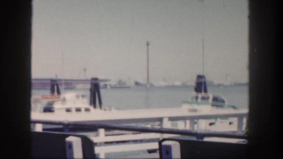 1962 San Diego pier view with a white docked boat, railings, bench backs, harbor water, and distant waterfront structures.