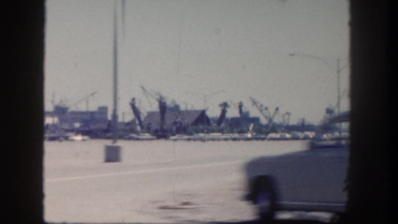A passing car blurs across a 1962 San Diego harbor roadside view with docked boats, cranes, and waterfront structures beyond.