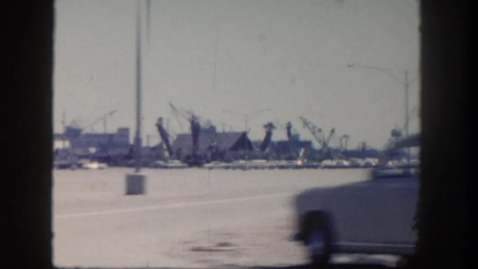 A passing car blurs across a 1962 San Diego harbor roadside view with docked boats, cranes, and waterfront structures beyond.