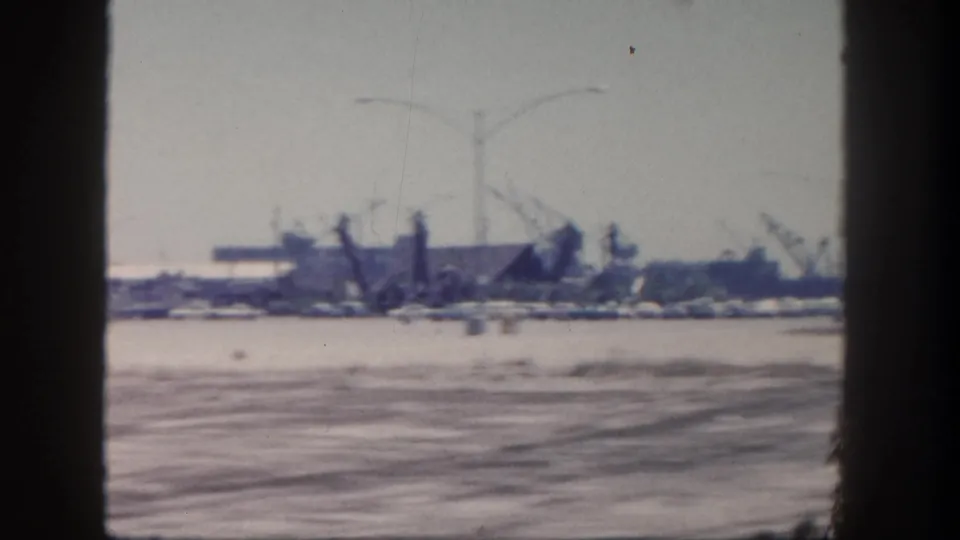 1962 San Diego harbor view with rippled water in the foreground and cranes, boats, streetlights, and low waterfront buildings beyond.