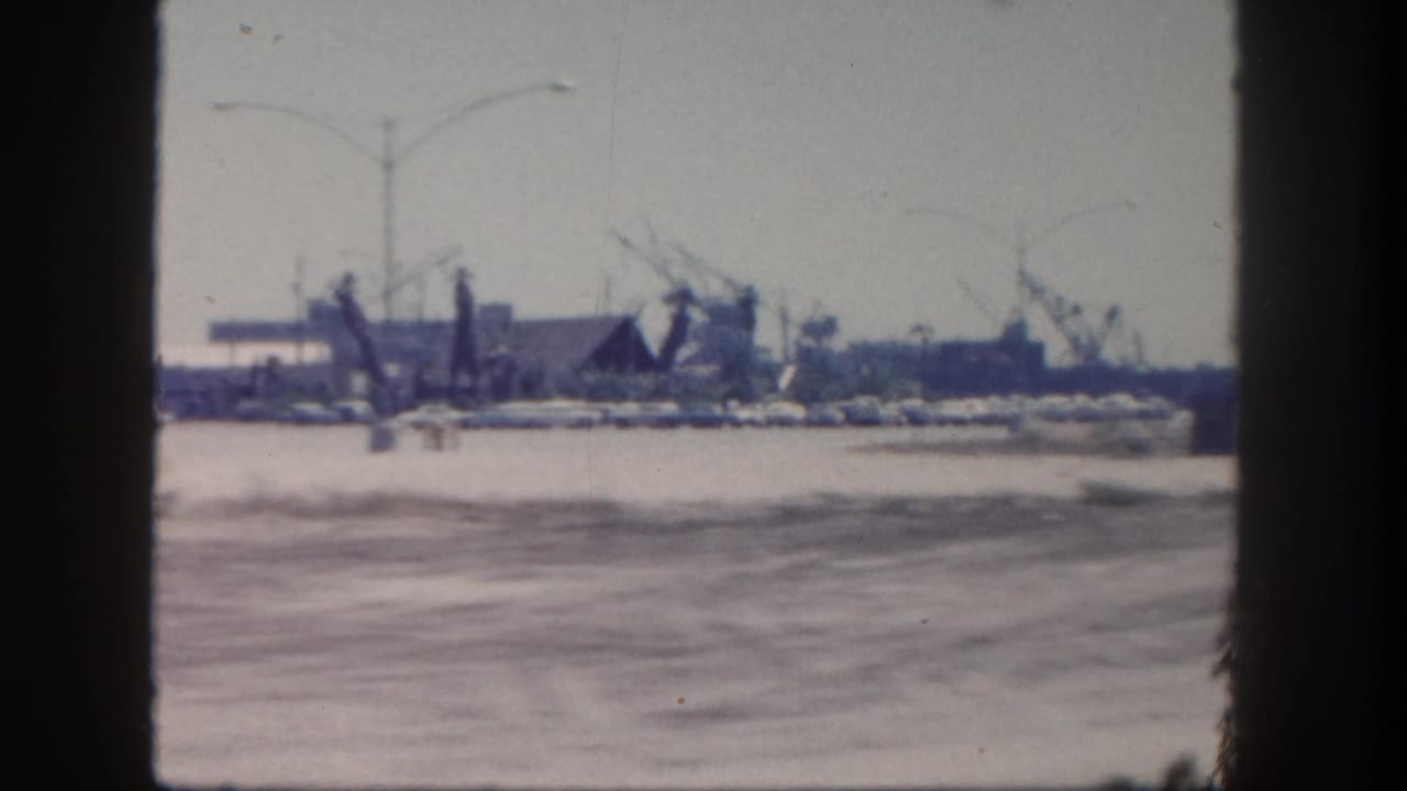 1962 San Diego harbor view from a moving vehicle, with a broad road foreground and distant fishing boats, masts, cranes, and waterfront buildings.