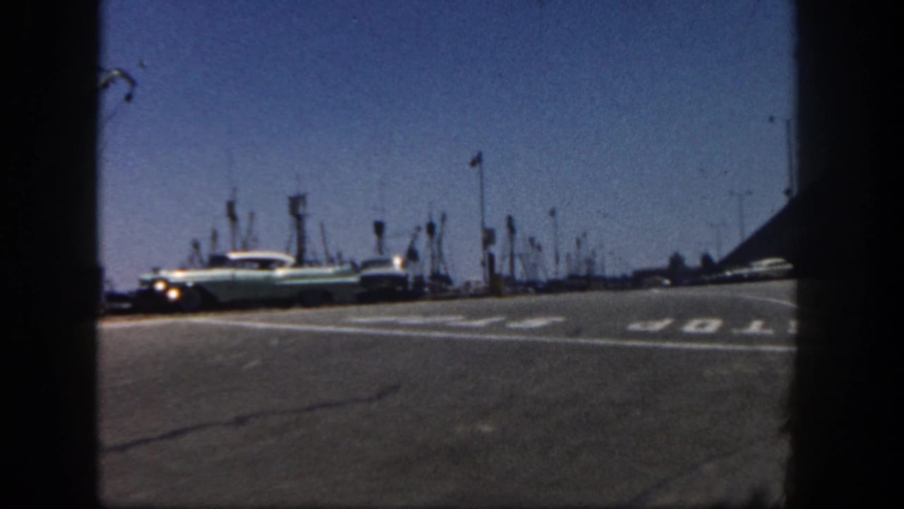 A light-colored 1960s sedan passes near a painted STOP marking on a San Diego harbor road, with fishing-boat masts and poles in the background.