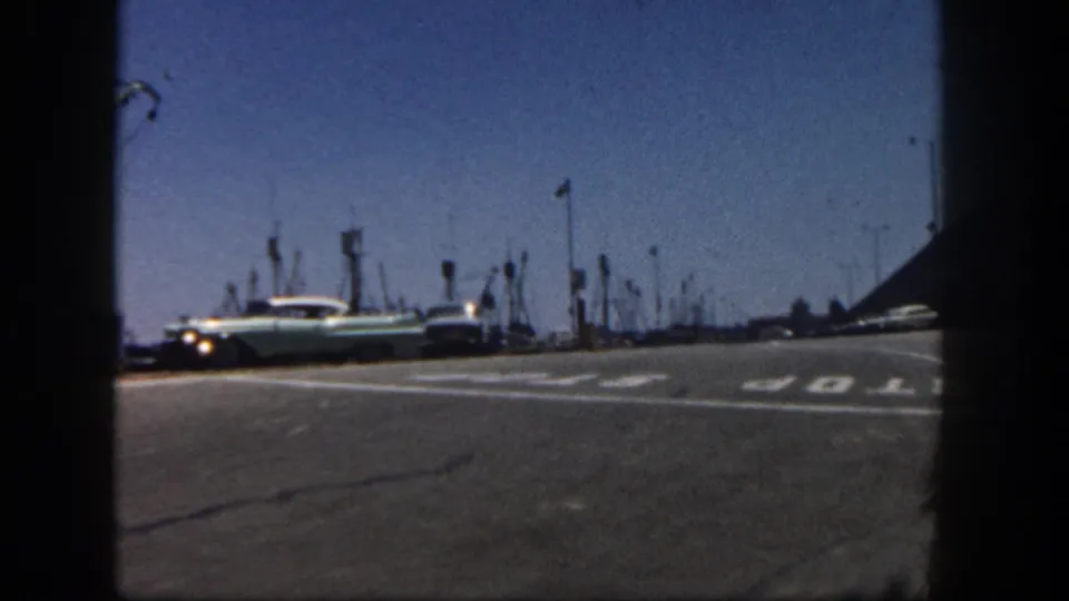 A light-colored 1960s sedan passes near a painted STOP marking on a San Diego harbor road, with fishing-boat masts and poles in the background.