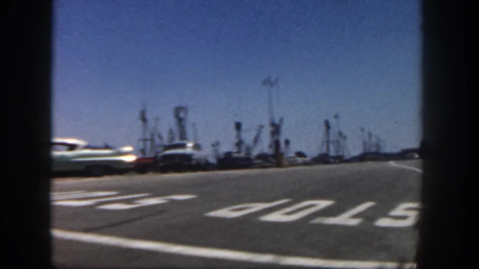 Moving-vehicle view of a San Diego harbor road with large STOP pavement lettering, a passing classic car, and clustered fishing-boat masts beyond.
