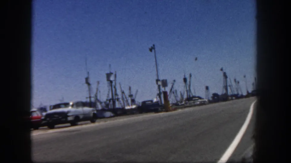 San Diego harbor road curves past classic cars and waterfront masts in 1962 home-movie footage, framed by dark 8mm film edges.