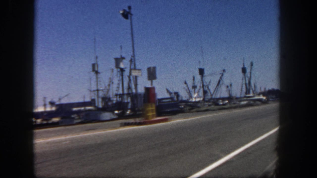 San Diego harbor from a moving 8mm home movie, showing docked fishing boats, masts, road surface, curb markers, and blue sky.