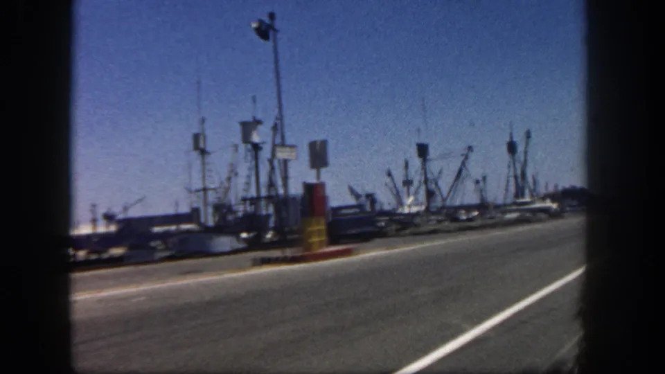 San Diego harbor from a moving 8mm home movie, showing docked fishing boats, masts, road surface, curb markers, and blue sky.