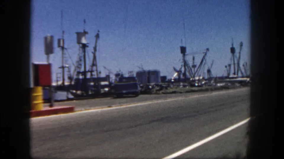 1962 San Diego waterfront road scene with a parked car, dockside posts, fishing boats, rigging, and soft 8mm motion blur.