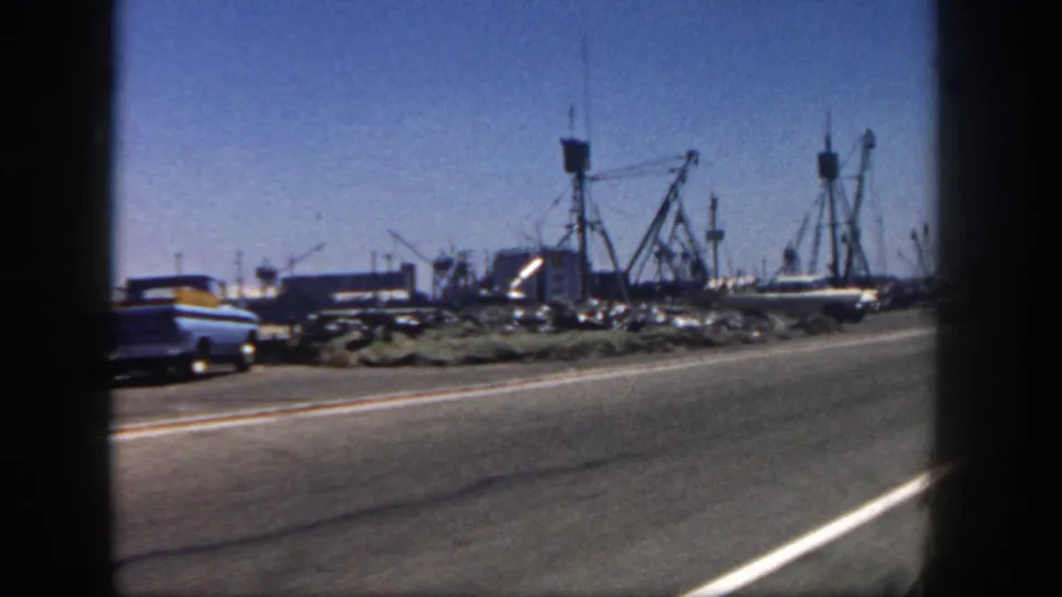 End of the 1962 San Diego harbor pass, with a blue car near the road, docked fishing boats, masts, rocks, and clear sky.