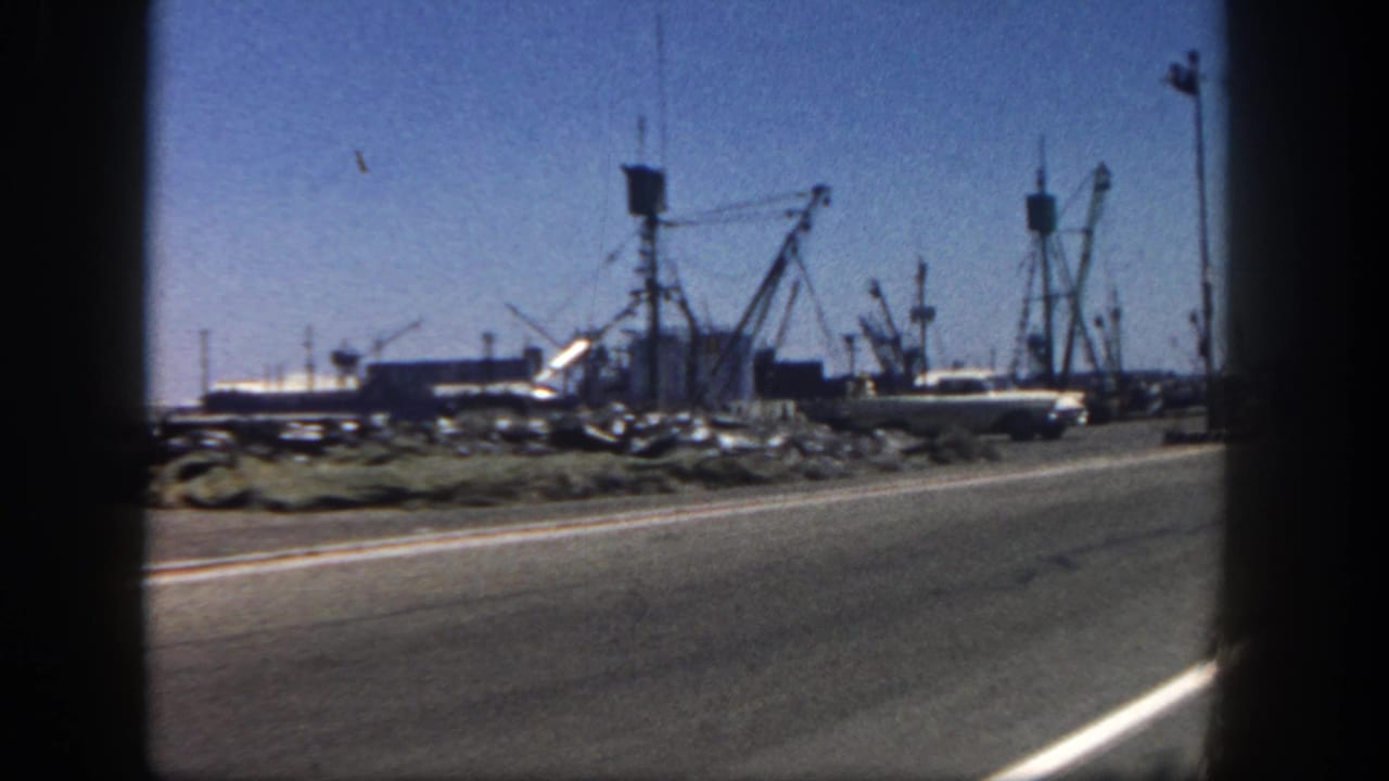 Harbor road with parked cars, boats, cranes, and dark film-frame edges.