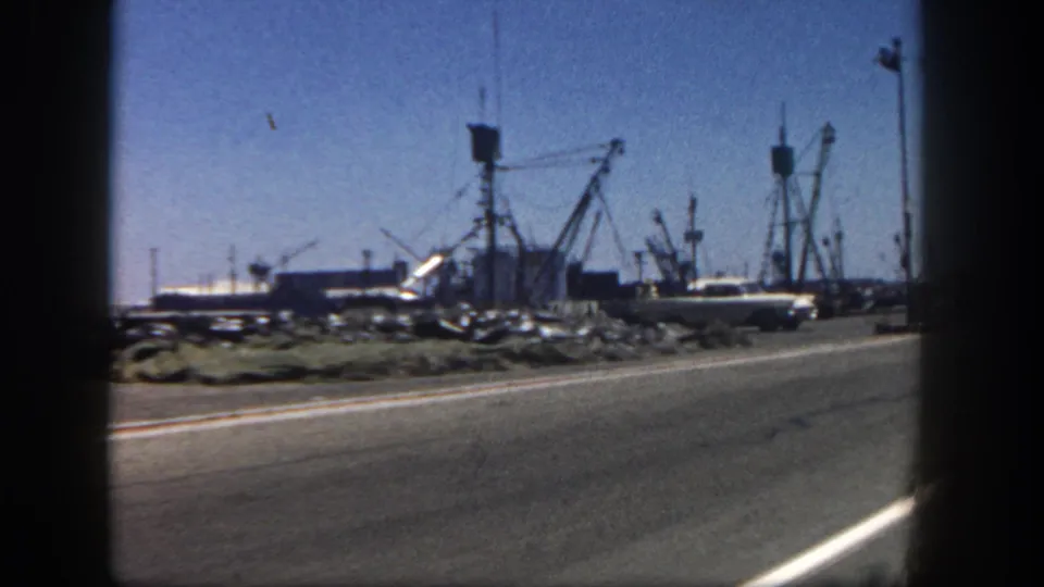 1962 San Diego harbor road view from a moving vehicle, with parked cars, fishing boats, dock cranes, and blue sky.