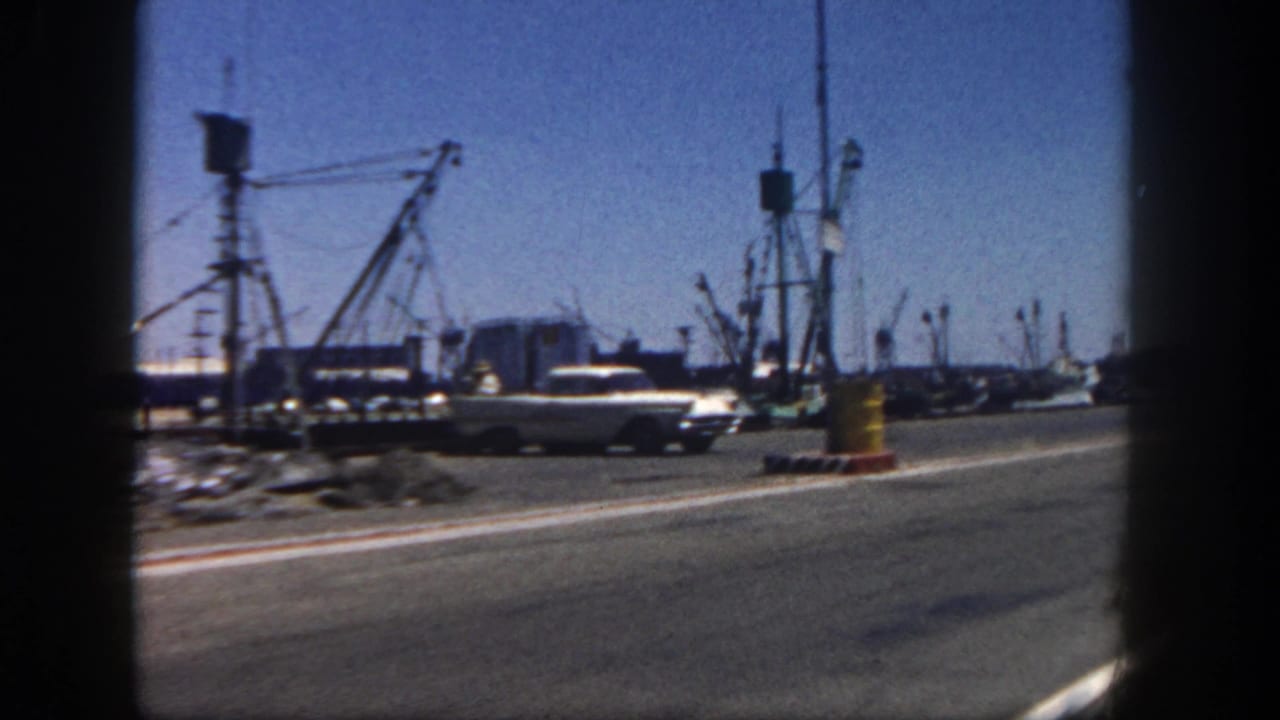 San Diego harbor roadside scene with early 1960s cars, boats, cranes, dock structures, and a yellow roadside barrel.
