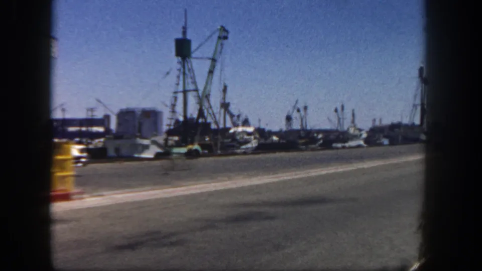 Passenger-side 8mm view of a San Diego harbor road, with cranes, boats, parked cars, and small dockside buildings.