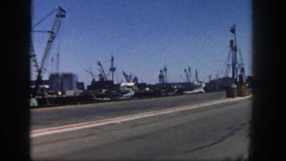 Wide moving-vehicle view along a 1962 San Diego harbor road with fishing boats, cranes, dock equipment, and strong 8mm gate shadows.