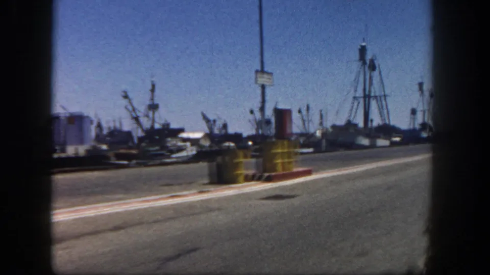 1962 San Diego harbor road scene showing yellow roadside barrels, moored boats, cranes, mast-like structures, and industrial waterfront equipment.