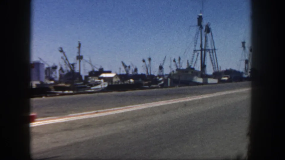 San Diego harbor drive-by in 1962, with a paved road in the foreground and boats, cranes, and tall dockside structures beyond it.