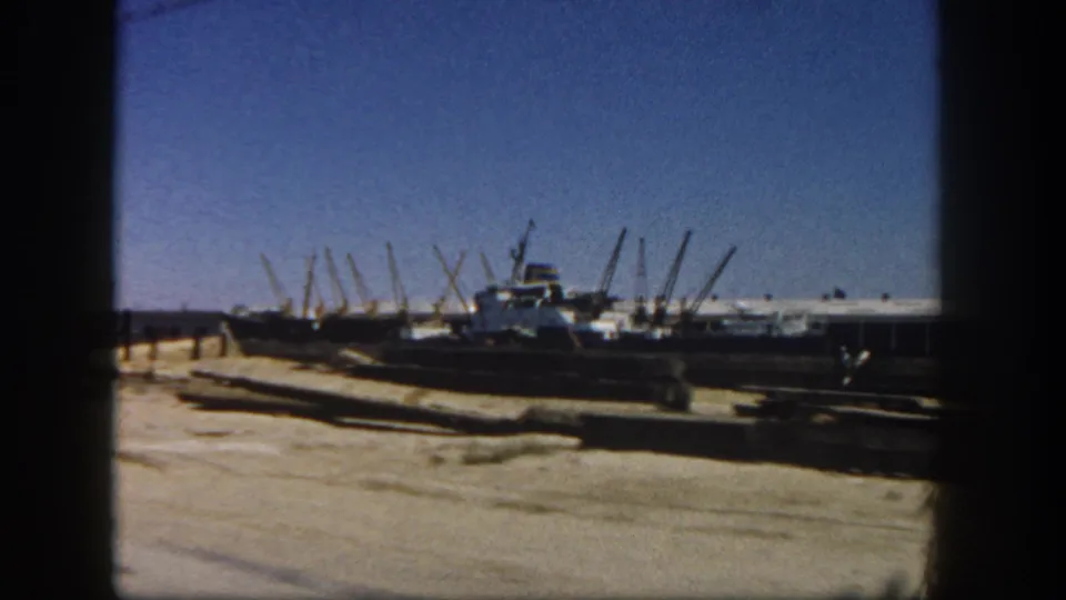 1962 8mm harbor frame showing dock pavement, moored boats, crane booms, low waterfront buildings, and a clear blue sky in San Diego.